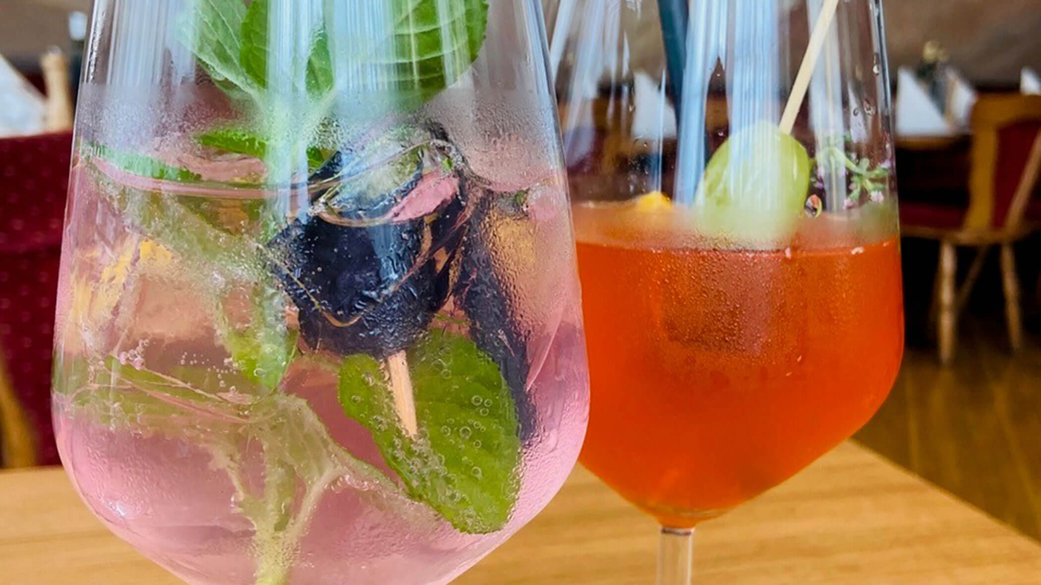 Two colorful cocktails in glasses, one pink with mint and berries, one orange with fruit garnish, on a table. - Il Leone Restaurant