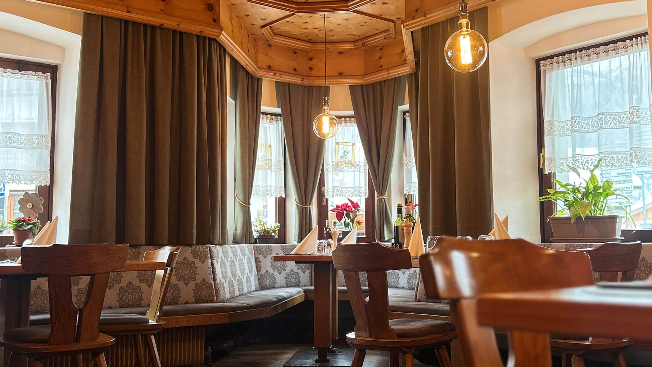 Cozy restaurant interior with wooden furniture, hanging lights, and large windows with curtains and potted plants. - Il Leone Restaurant