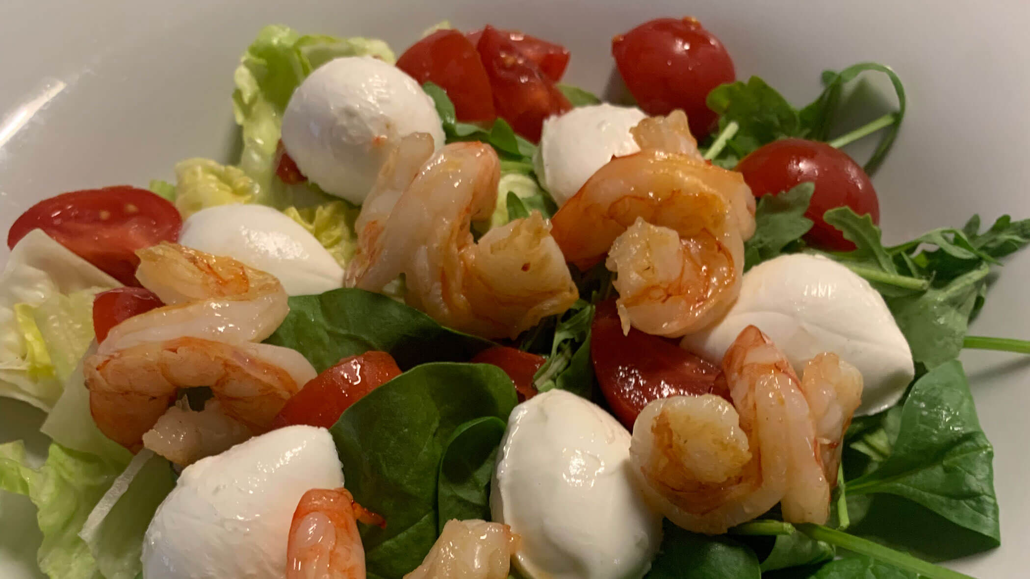 Salad with shrimp, mozzarella balls, cherry tomatoes, and leafy greens in a white bowl. - Il Leone Restaurant