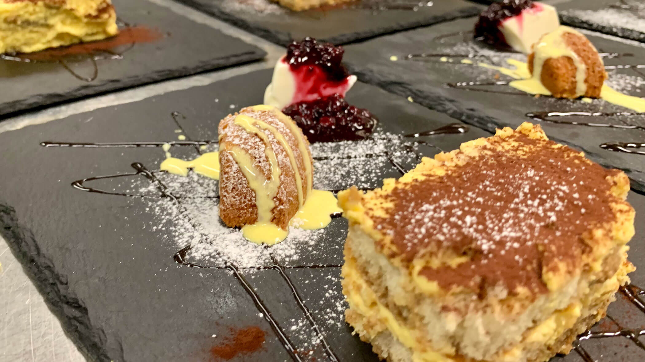 Close-up of tiramisu slices and pastries garnished with sauce and powdered sugar on slate plates. - Il Leone Restaurant