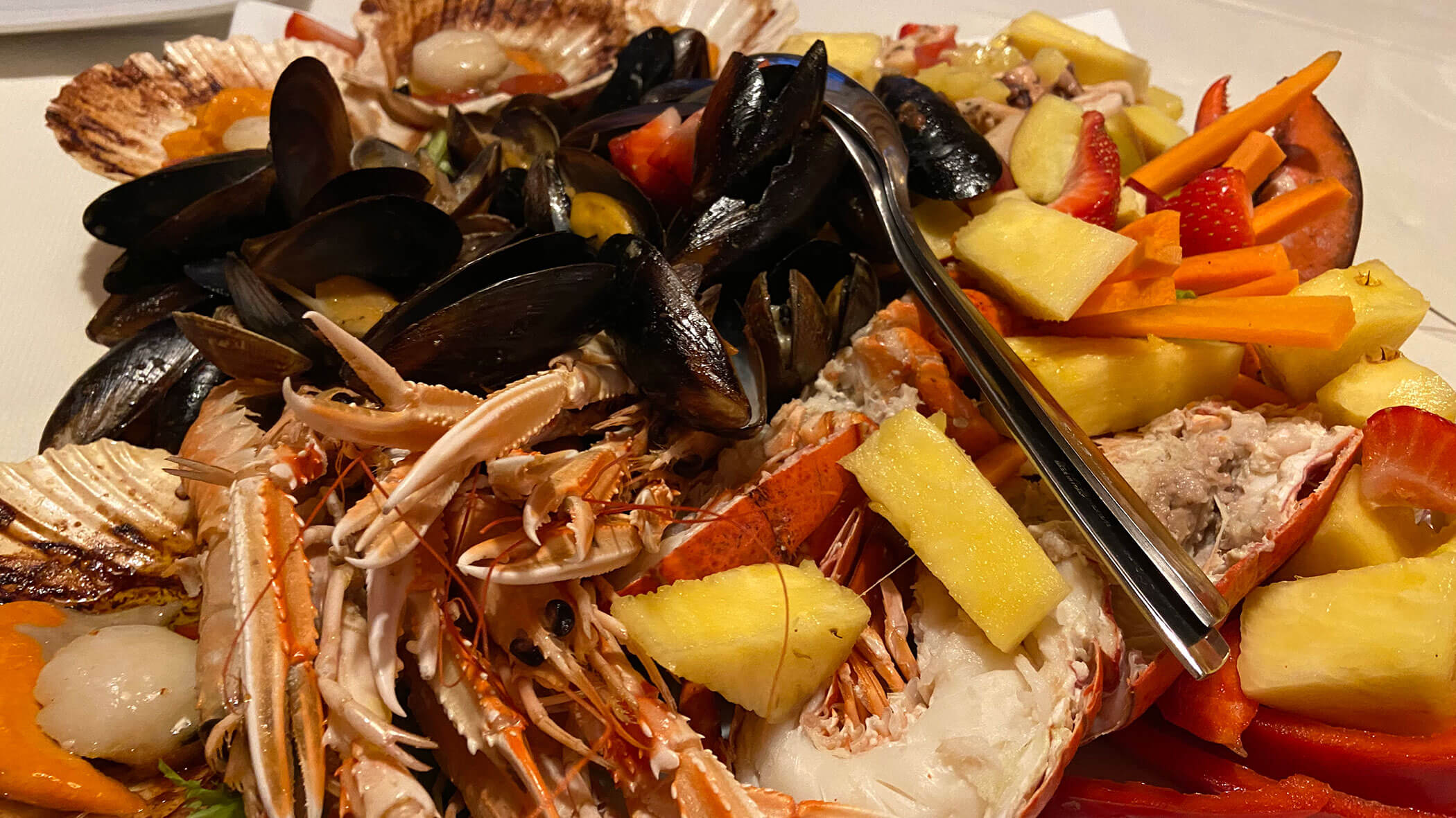 A plate of assorted seafood, mussels, lobster, scallops, and fresh fruit slices with a fork. - Il Leone Restaurant