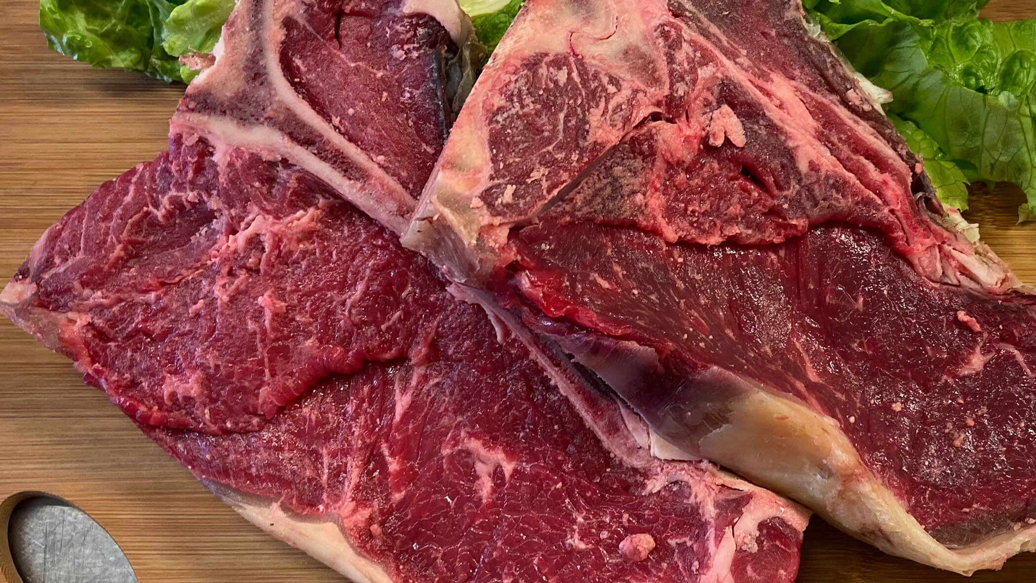 Two raw T-bone steaks on a wooden board, with green lettuce leaves beside them. - Il Leone Restaurant