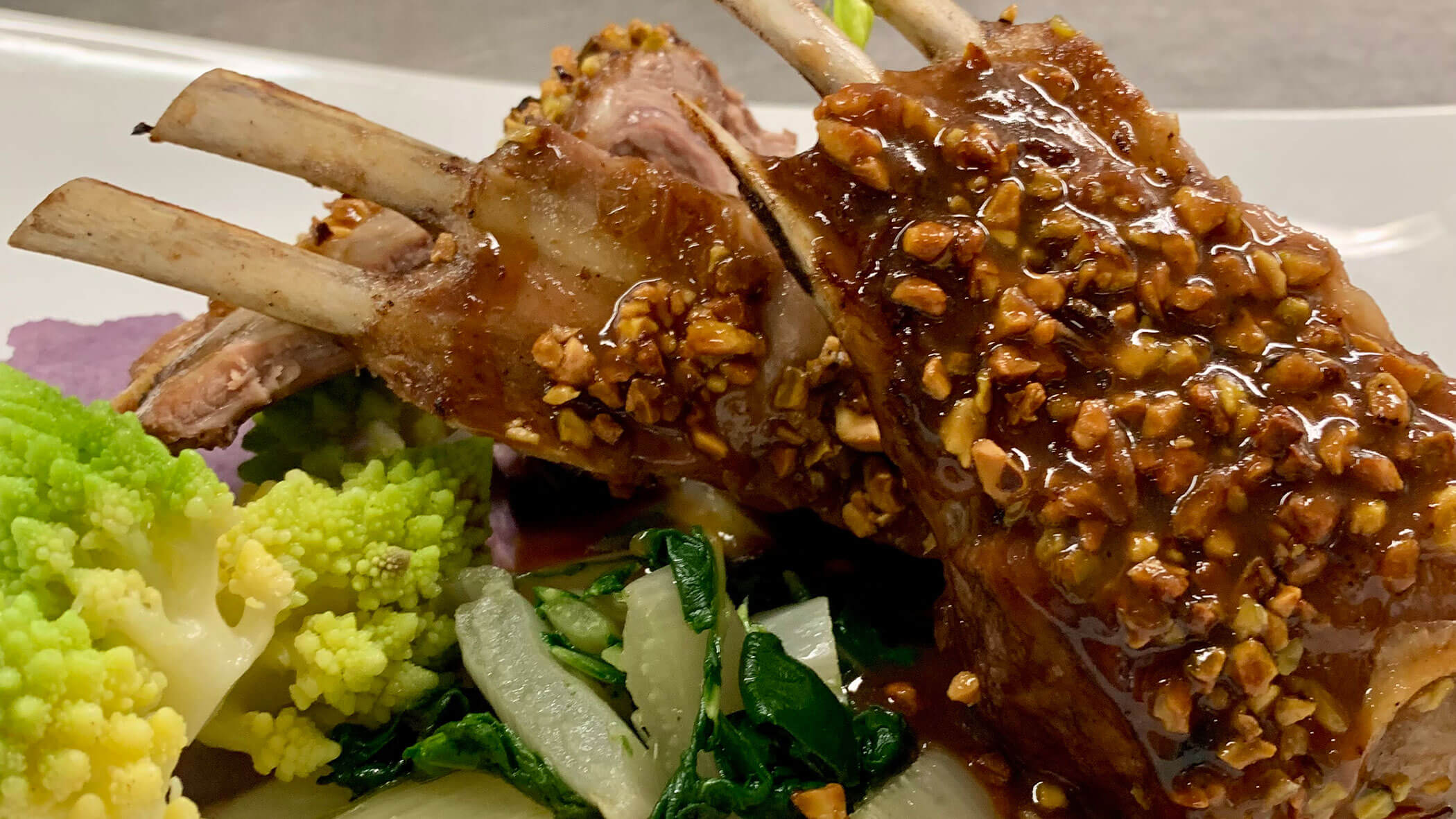 Rack of lamb with a nutty glaze, served with mixed sautéed greens and Romanesco broccoli on a white plate. - Il Leone Restaurant