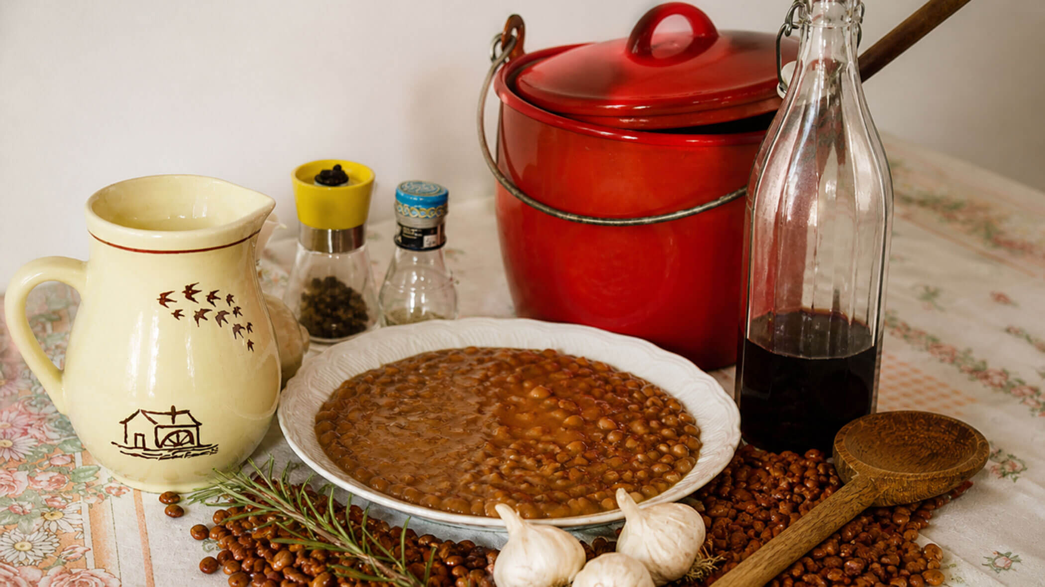 A plate of bean stew with garlic, herbs, wine, condiments, and a red pot on a table. - Il Leone Restaurant