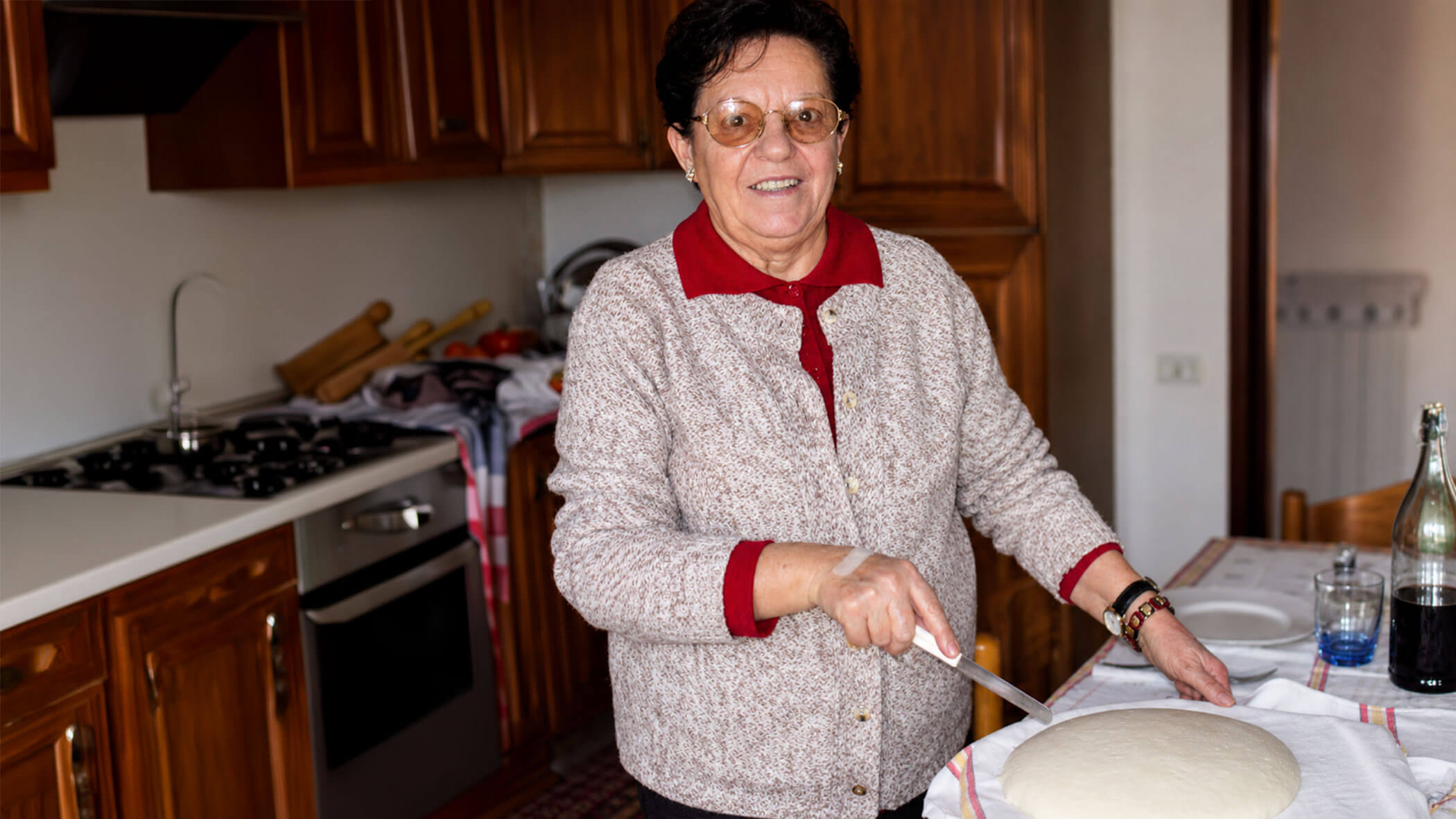 Smiling elderly woman stands in a kitchen, cutting dough on a table covered with a white cloth. - Il Leone Restaurant