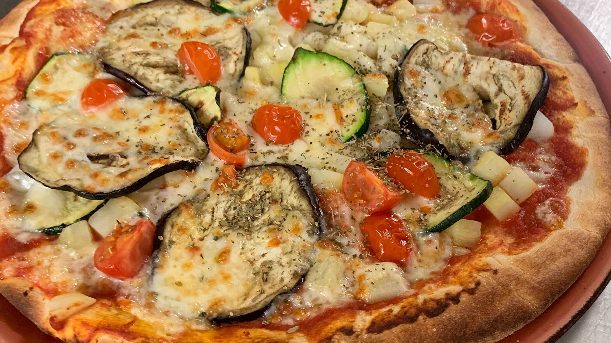 A pizza topped with eggplant, zucchini, cherry tomatoes, cheese, and herbs on a tomato sauce base. - Il Leone Restaurant